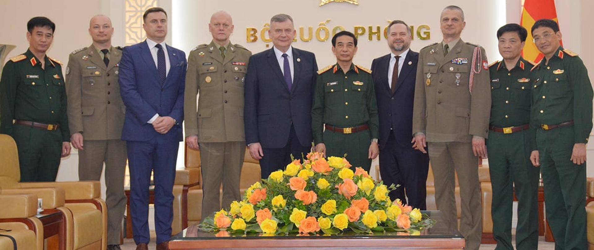 Wiceminister Konrad Gołota, wiceminister obrony narodowej Paweł Bejda, przedstawiciele PGZ S.A., PIT-RADWAR oraz Socjalistycznej Republiki Wietnamu stoją na tle zdobionej ściany z napisami. Przed nimi kwiaty. 