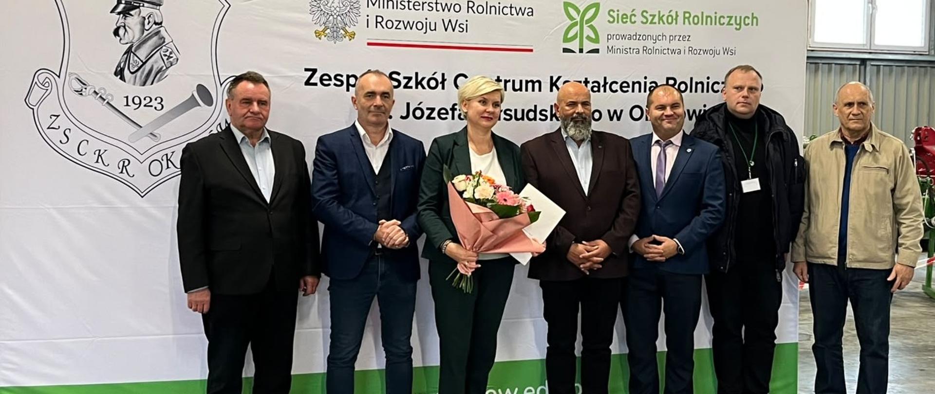 Uczestnicy wydarzenia pozują do zdjęcia grupowego przed banerem Zespołu Szkół Centrum Kształcenia Rolniczego w Okszowie.