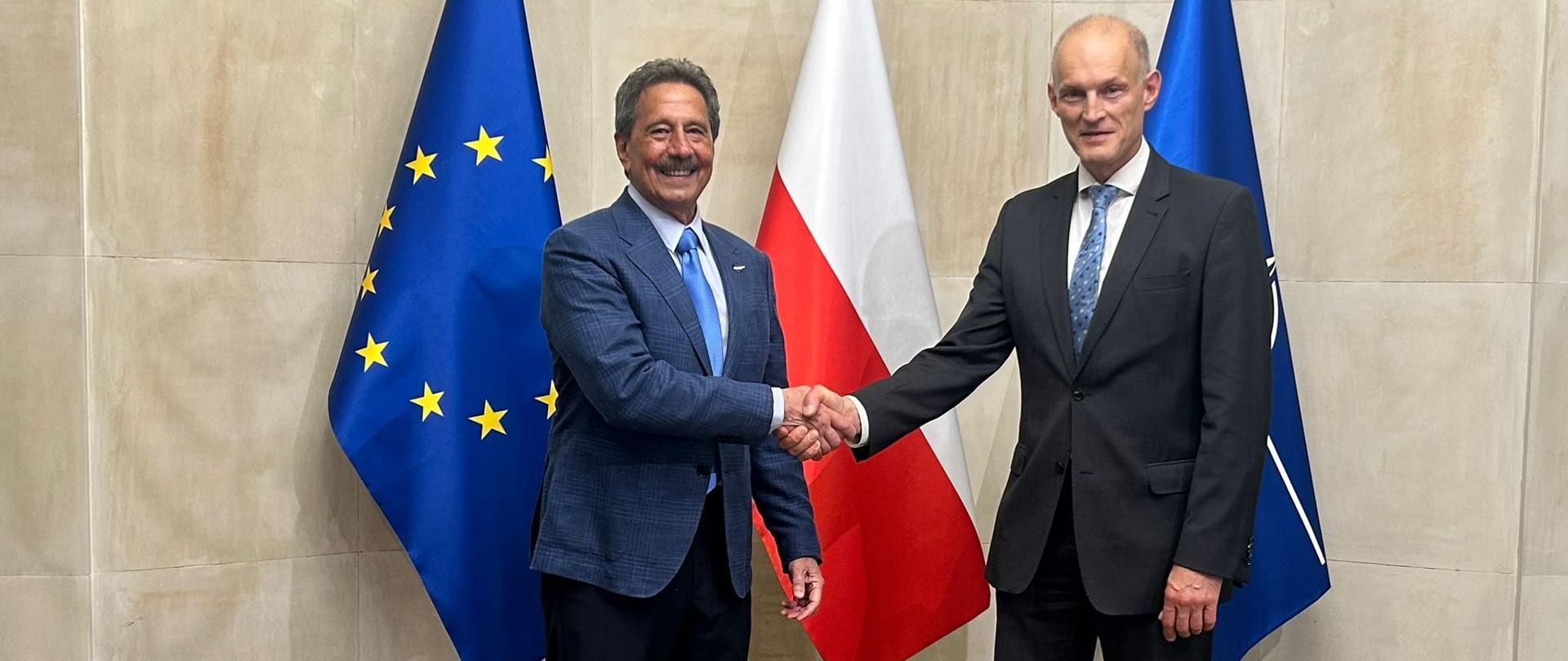 Podsekretarz stanu Wojciech Zajączkowski podczas uścisku dłoni z ministrem Hondurasu. W tle trzy flagi.