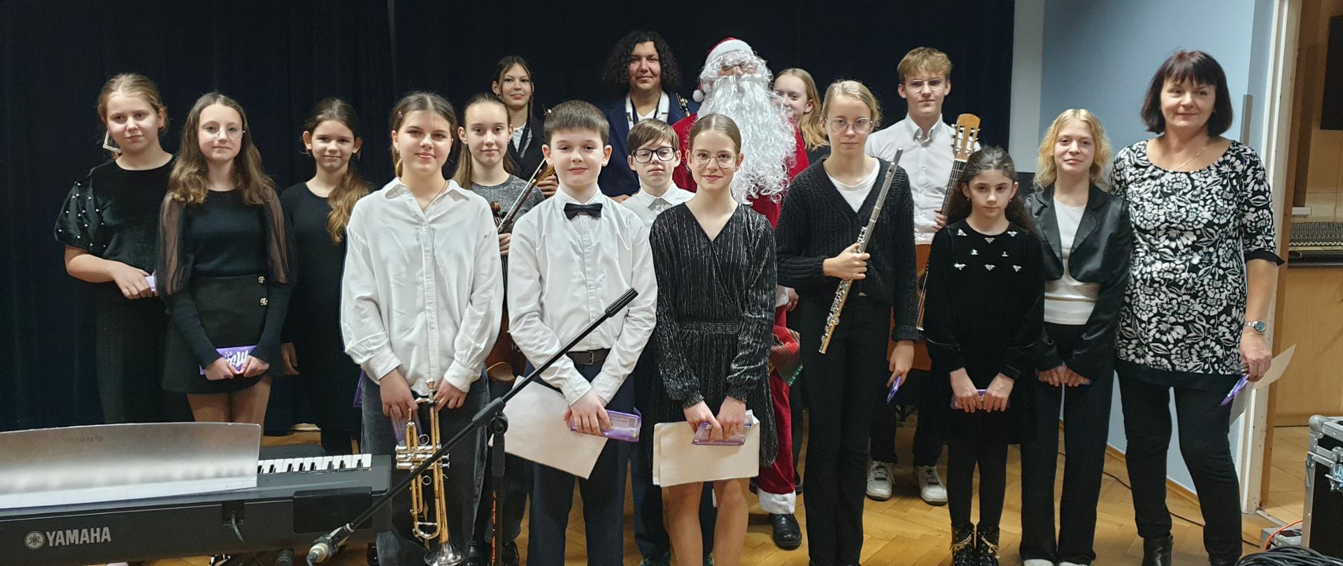 Zdjęcie przedstawia grupę młodzieży i kilku dorosłych po koncercie. Uczestnicy są elegancko ubrani – większość w strojach galowych lub wieczorowych, w odcieniach czerni i bieli. Na pierwszym planie znajduje się keyboard Yamaha i mikrofon. Część uczniów trzyma instrumenty, takie jak trąbka, skrzypce, flet poprzeczny i gitara klasyczna. W centrum zdjęcia znajduje się postać Świętego Mikołaja, co podkreśla świąteczny charakter wydarzenia. Grupa stoi na tle ciemnej, aksamitnej kurtyny scenicznej, a po prawej stronie widać otwarte drzwi prowadzące do innego pomieszczenia. 