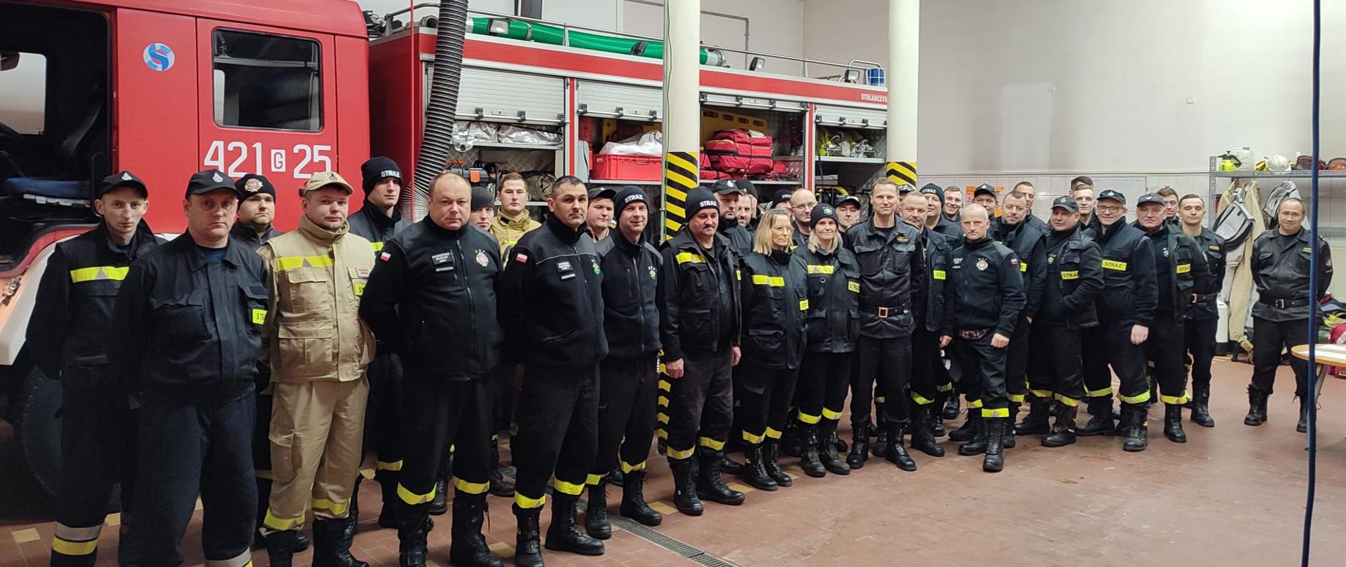 W garażu Jednostki Ratowniczo-Gaśniczej w Człuchowie stoją w dwuszeregu uczestnicy szkolenia. Wśród nich Komendant Powiatowy Państwowej Straży Pożarnej w Człuchowie st. bryg. Ernest Szczepański oraz funkcjonariusze będący w składzie komisji egzaminacyjnej. W tle samochód pożarniczy.