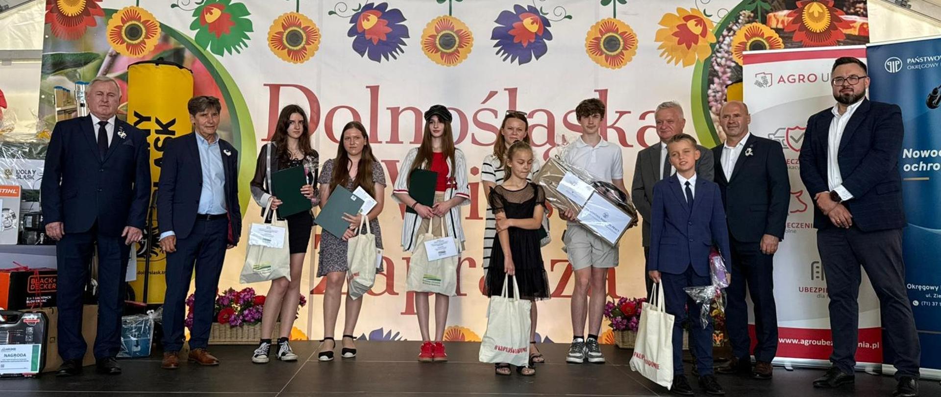 Grupa siedmiorga dzieci pozuje do zdjęcia na scenie w towarzystwie pięciu mężczyzn w garniturach. Dzieci trzymają w rękach dyplomy i torby z nagrodami.