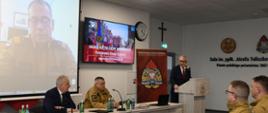 Mężczyzna stoi przy mównicy i przemawia do funkcjonariuszy którzy siedzą na krzesłach na ekranach wyświetlana jest prezentacja.