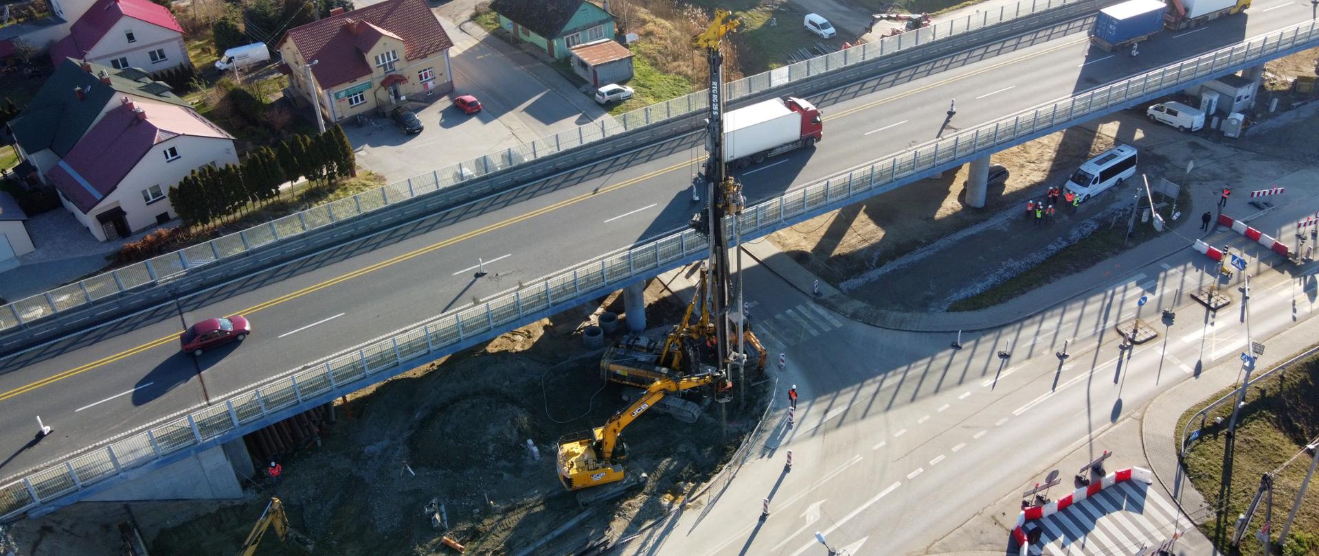 Świeci słońce. Widok z góry na pracę palownicy w miejscowości Nienadówka, gdzie powstaje nowy obiekt. Z lewej strony widać domy, obok jest estakada, na której jadą auta. A po prawej stronie istniejącej estakady pracuje palownica.