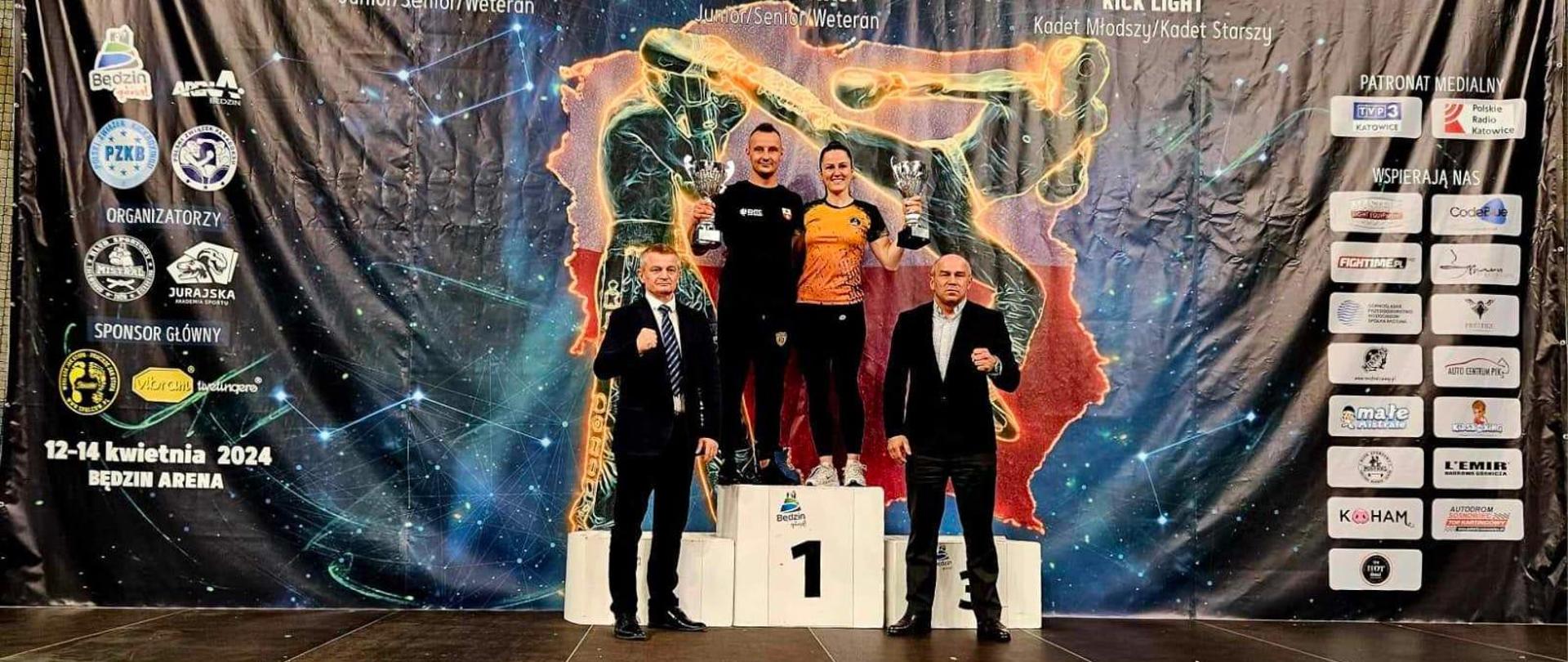 Strażak z Elbląga stoi na najwyższym miejscu podium, w ręku trzyma puchar. Za plecami wielki baner z napisem Mistrzostwa Polski w kickboxingu.