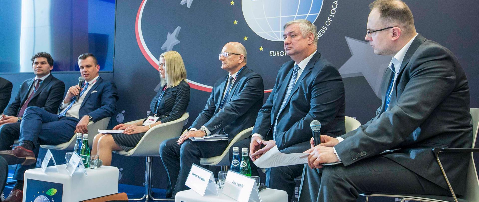 Na zdjęciu uczestnicy debaty - 6 osób siedzących w fotelach. Wiceminister Adam Hamryszczak trzyma w ręku mikrofon i mówi.