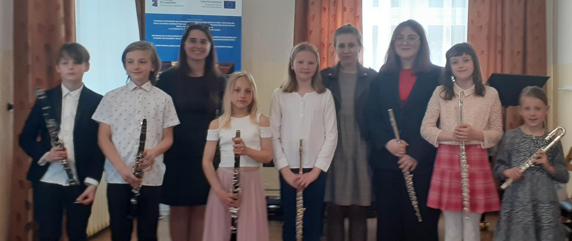 Wszyscy uczestnicy audycji wraz z nauczycielkami stoją w rzędzie . Uczniowie trzymają swoje instrumenty w pozycji pionowej. Nad uczestnikami wydarzenia świecą wiszące cztery lamy. Za nimi dwa okna przysłonięte firankami i zasłonami okna, pulpit oraz baner unijny.