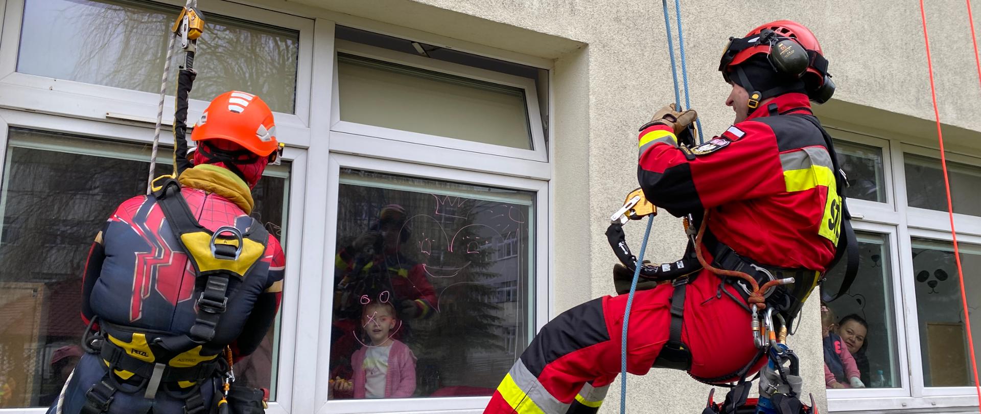 Zdjęcie przedstawia strażaka oraz alpinistę opuszczonych na linach za pomocą technik alpinistycznych do okna szpitala przez, które machają do dzieci znajdujących się w środku.
