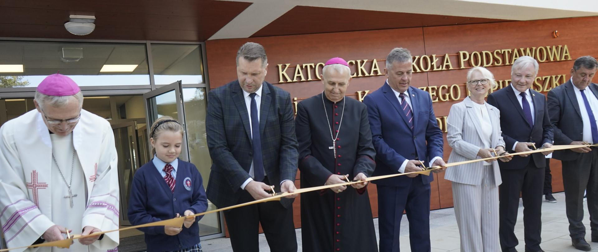 Grupa kilku osób stoi przed żółtą wstęgą, każdy trzyma nożyczki i równocześnie wszyscy przecinają wstęgę.