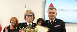 Funkcjonariusze Państwowej Straży Pożarnej stoją obok siebie jeden z nich trzyma obraz oraz kwiaty. 