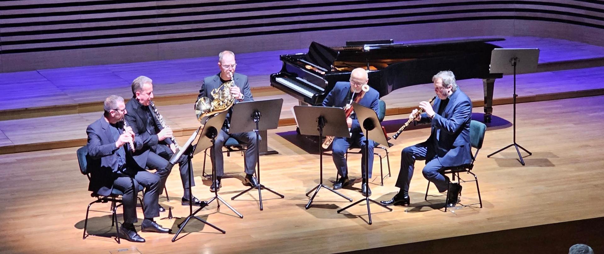 Pięciu mężczyzn gra na instrumentach dętych: flecie, klarnecie, waltorni, fagocie i oboju.