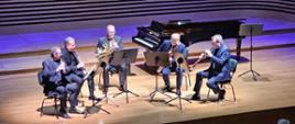Pięciu mężczyzn gra na instrumentach dętych: flecie, klarnecie, waltorni, fagocie i oboju.