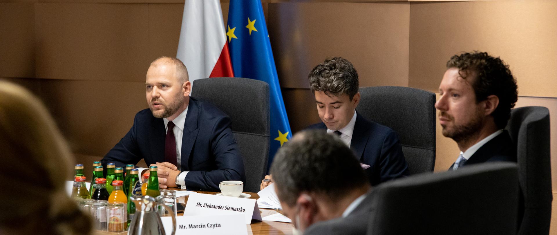 Wiceminister Krzysztof Mazur siedzi za stołem na tle flag polskiej i unijnej. Obok delegacja Polski.