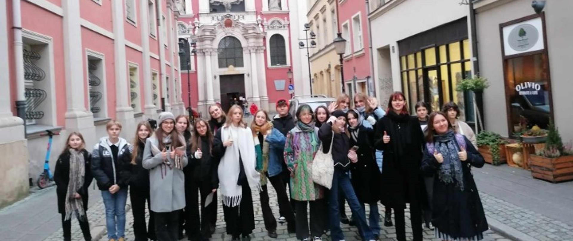 Zdjęcie kolorowe. Grupa uczniów stojących na brukowanej ulicy. W tle barokowa fasada kościoła.