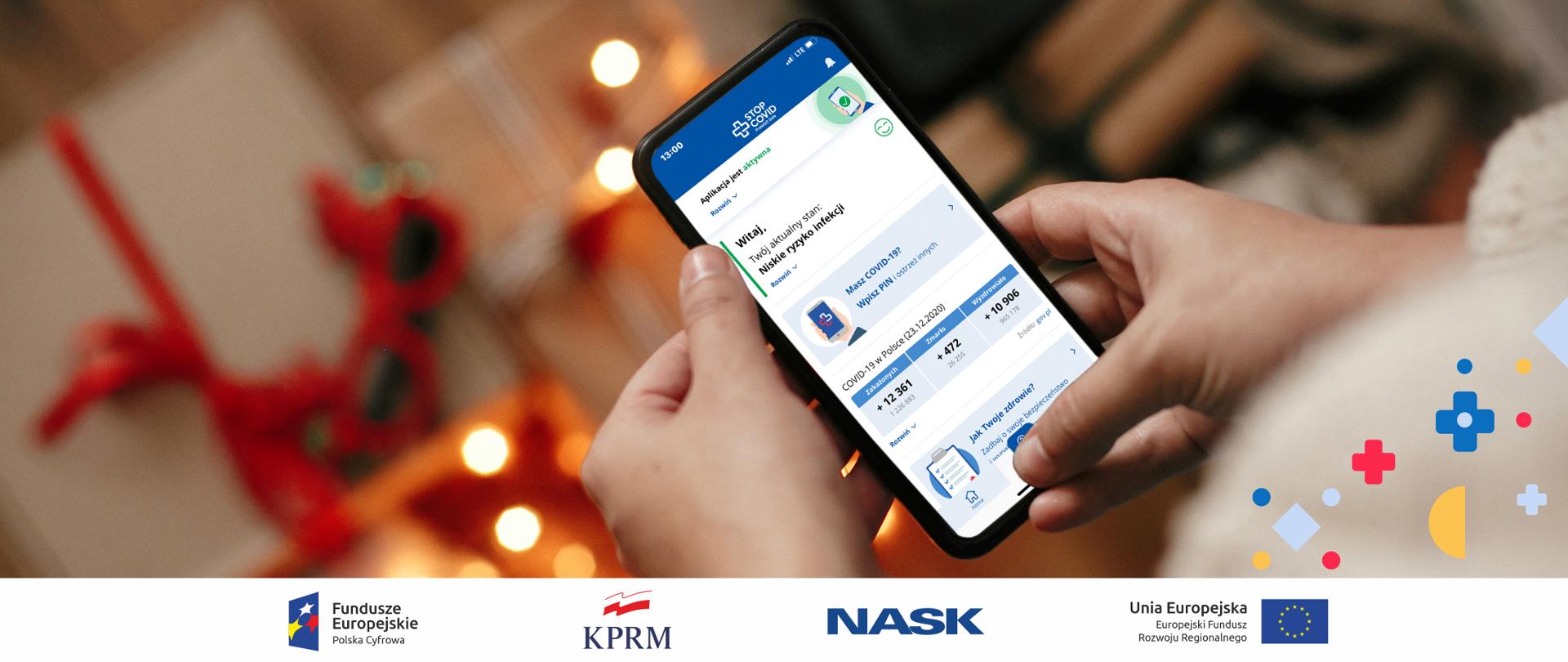 Widok na dłonie trzymające smartfona, na jego ekranie wyświetla się aplikacja STOP COVID - ProteGo Safe. W tle, na drugim mniej wyraźnym planie, widoczne świąteczne ozdoby, w tym m.in. lampki.