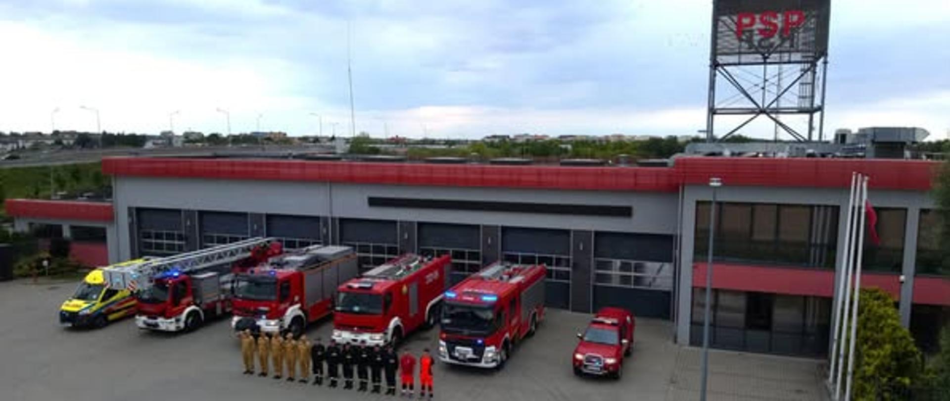Zdjęcie zostało wykonane przed Jednostką Straży Pożarnej w Radomiu. Funkcjonariusze oraz ratownicy medyczni ustawieni są w pozycji zasadniczej - na baczność. Za funkcjonariuszami ustawione są wozy strażackie oraz jeden samochód Zespołu Ratownictwa Medycznego. Funkcjonariusze ubrani są w służbowe mundury koloru żółtego oraz czarnego.
