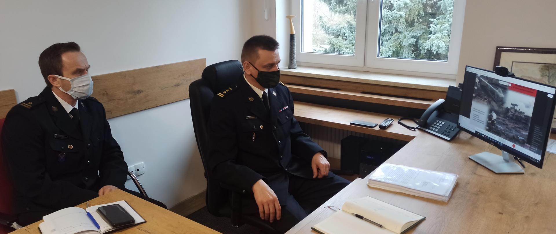 Zdjęcie przedstawia Komendanta Powiatowego PSP w Brodnicy oraz Dowódcę JRG podczas przeprowadzonej w trybie zdalnym narady rocznej kadry kierowniczej Państwowej Straży Pożarnej w województwie kujawsko-pomorskim. Komendant i Dowódca siedzący przy biurku w mundurze oglądają prezentowane materiały.