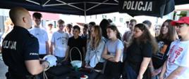 Policjant w mundurze prezentuje wyposażenie służbowe grupie młodzieży. Na stole wystawione są m.in. kajdanki, pałka, kask oraz tarcza do zatrzymywania pojazdów.