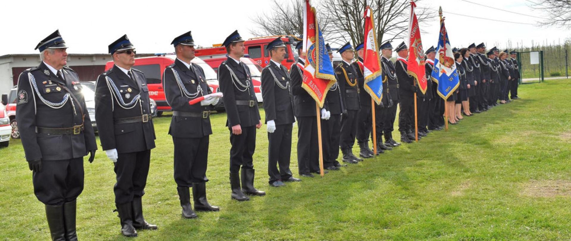 Na zdjęciu widoczne poczty sztandarowe, poczet flagowy oraz dowódcę pododdziału, stojące w jednym szeregu na murawie trawiastej. Na drugim planie samochody strażackie.