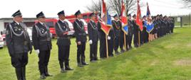 Na zdjęciu widoczne poczty sztandarowe, poczet flagowy oraz dowódcę pododdziału, stojące w jednym szeregu na murawie trawiastej. Na drugim planie samochody strażackie.