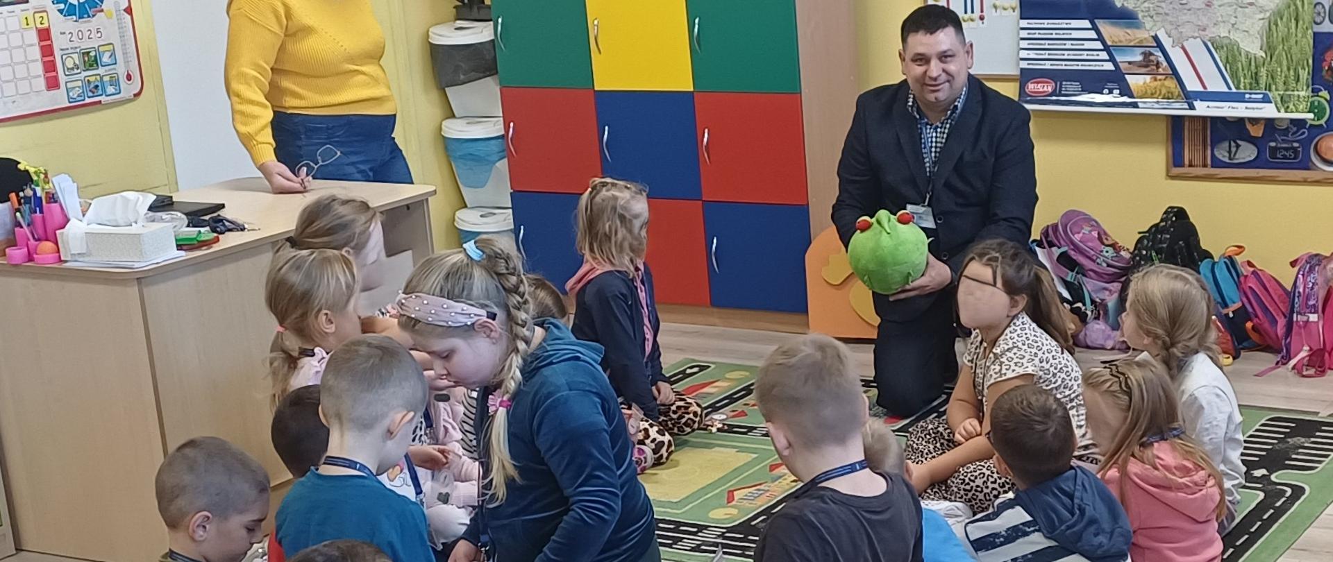 Grupa dzieci siedząca na dywanie w kolorowej Sali. Wśród nich kuca mężczyzna ubrany czarną marynarkę i trzyma w dłoniach zieloną maskotkę. W tle widać kolorowe szafki. Po lewej stronie przy biurku stoi kobieta w żółtym swetrze. 
