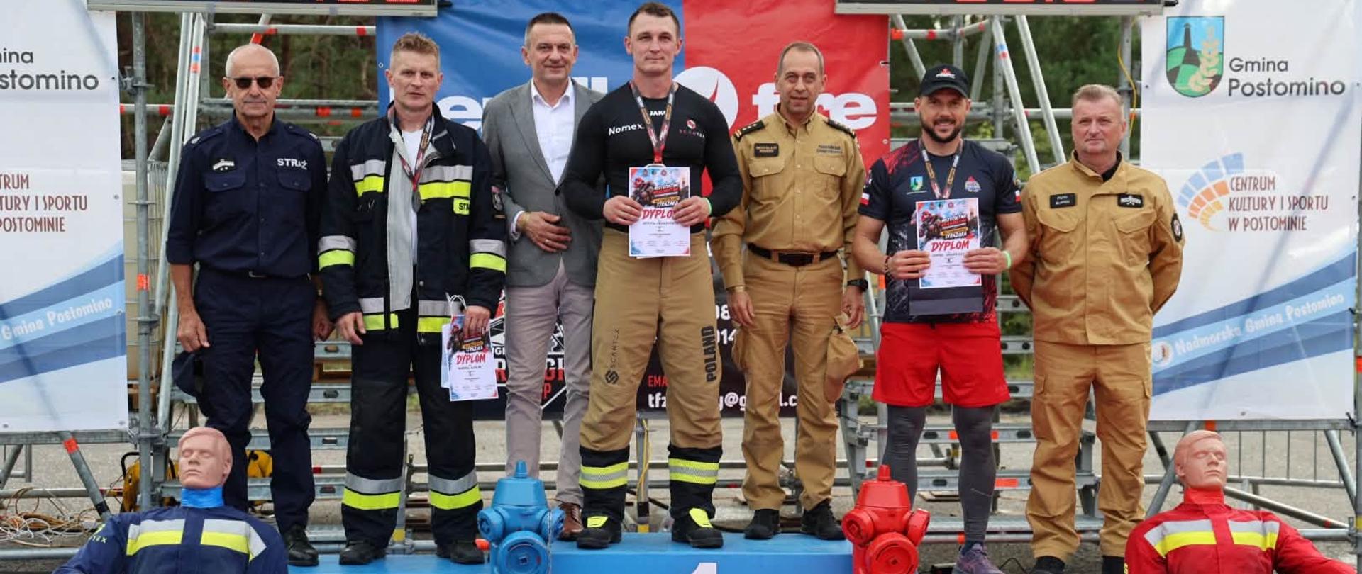 Dekoracja zwycięzców Mistrzostw Województwa Zachodniopomorskiego Najtwardszego Strażaka w Jarosławcu. Na podium stoją nagrodzeni zawodnicy oraz organizatorzy wydarzenia.