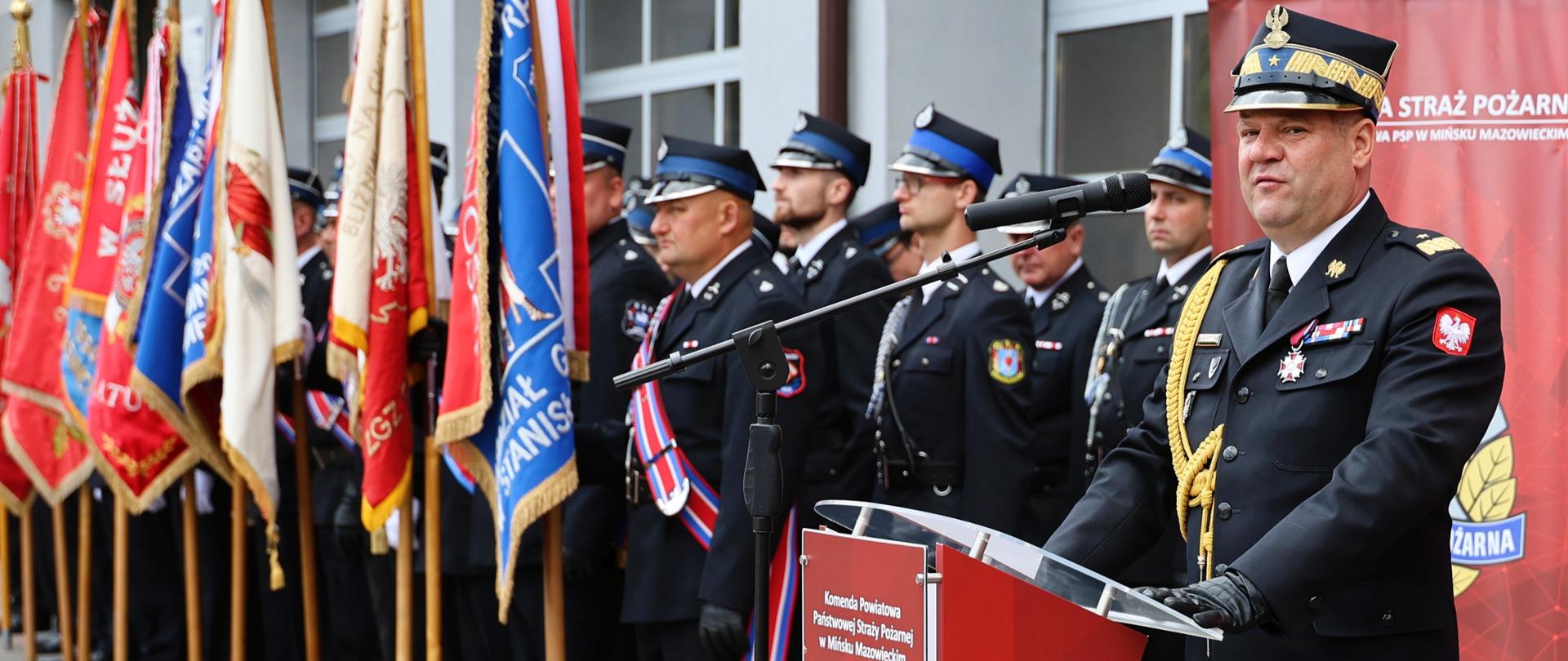  Ujęcie z boku na nadbryg. Artura Gonerę stojącego za oficjalną czerwoną mównicą z logo Komendy Powiatowej Państwowej Straży Pożarnej w Mińsku Mazowieckim. Komendant przemawia do mikrofonu, patrząc przed siebie. Po lewej stronie, wzdłuż placu, stoi poczet sztandarowy prezentujący szereg różnobarwnych sztandarów formacji strażackich. Za komendantem, w tle, widać rzędy strażaków stojących na baczność podczas oficjalnych przemówień.
