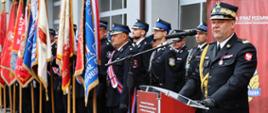  Ujęcie z boku na nadbryg. Artura Gonerę stojącego za oficjalną czerwoną mównicą z logo Komendy Powiatowej Państwowej Straży Pożarnej w Mińsku Mazowieckim. Komendant przemawia do mikrofonu, patrząc przed siebie. Po lewej stronie, wzdłuż placu, stoi poczet sztandarowy prezentujący szereg różnobarwnych sztandarów formacji strażackich. Za komendantem, w tle, widać rzędy strażaków stojących na baczność podczas oficjalnych przemówień. 