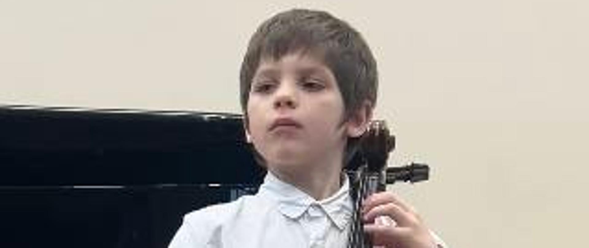 Uczeń Paweł Szczepaniak gra w stroju galowym na wiolonczeli na szkolnej sali koncertowej. 