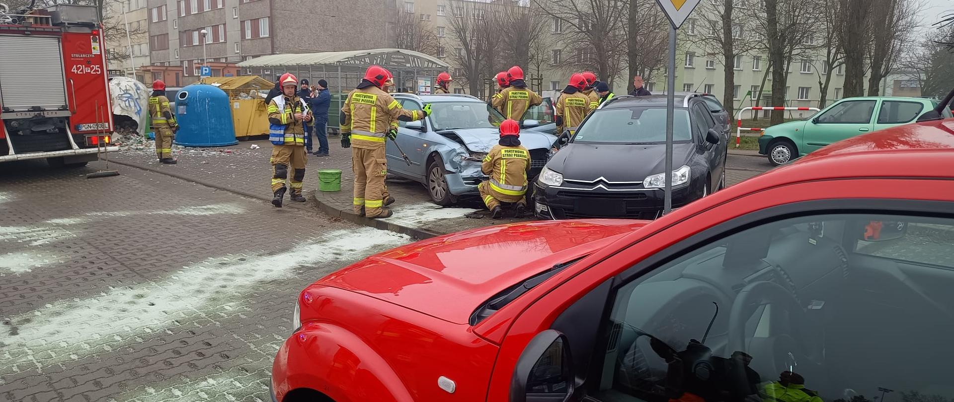 Na zdjęciu widać uszkodzone pojazdy przy których działają strażacy. Jest pochmurno, w tle widać bloki mieszkalne. Na jezdni widać rozsypany sorbent, który służy do zebrania rozlanych płynów eksploatacyjnych. 