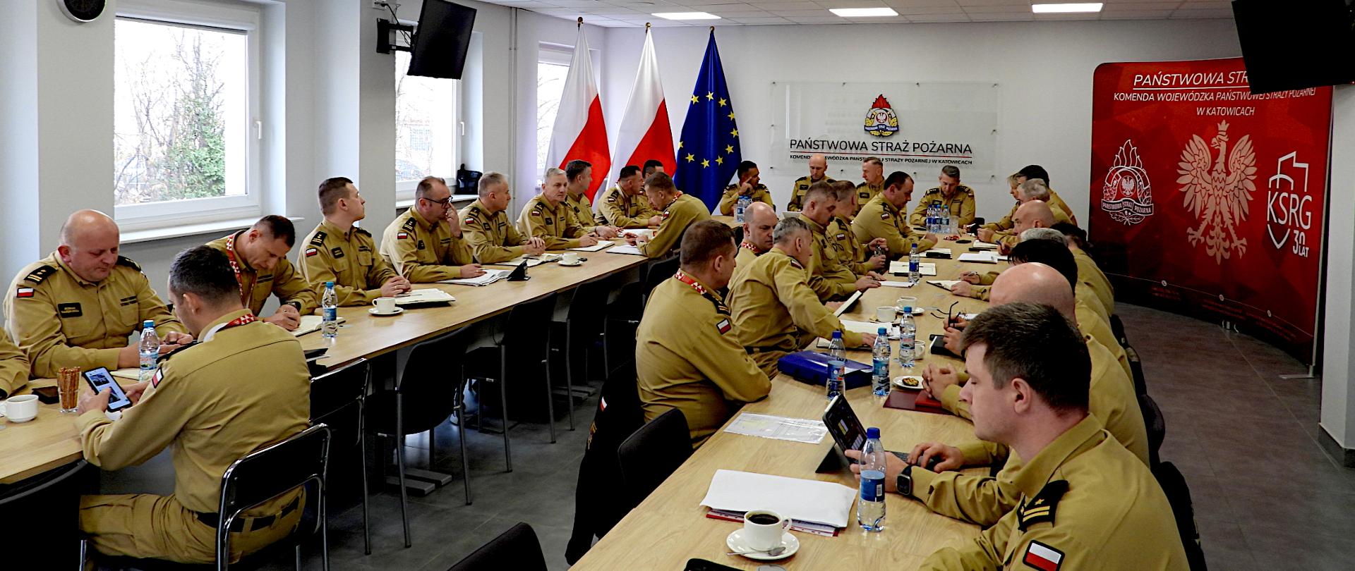 Na zdjęciu widać dużą salę sztabową w Komendzie Wojewódzkiej Państwowej Straży Pożarnej w Katowicach, w której przy długich stołach siedzi kilkudziesięciu mężczyzn w jednakowych, beżowych mundurach służbowych. To funkcjonariusze Państwowej Straży Pożarnej. Na ramionach mają naszywki z polską flagą. Wszyscy są skupieni — niektórzy piszą notatki, inni patrzą w dokumenty, tablety lub telefony. Na stołach stoją butelki z wodą, filiżanki z kawą i notatniki. W tle widoczne są trzy flagi: dwie biało-czerwone (Polski) i jedna niebieska z żółtymi gwiazdami (flaga Unii Europejskiej). Na ścianie znajduje się napis: „Państwowa Straż Pożarna, Komenda Wojewódzka Państwowej Straży Pożarnej w Katowicach”. Po prawej stronie stoi czerwone tło z logotypami PSP, godłem Polski i napisem „KSRG 30 lat”. Cała scena przedstawia naradę lub odprawę kierownictwa straży pożarnej.