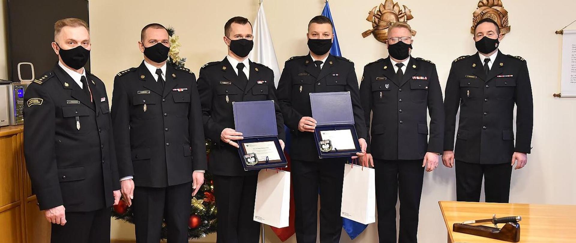 na zdjęciu sześciu strażaków w mundurach wyjściowych, stojących w pomieszczeniu, mężczyźni stoją w lekkim łuku, dwóch środkowych trzyma w rękach tabliczki okolicznościowe w niebieskich etui, za strażacki flagi Polski i UE, choinka oraz wyposażenie pomieszczenia