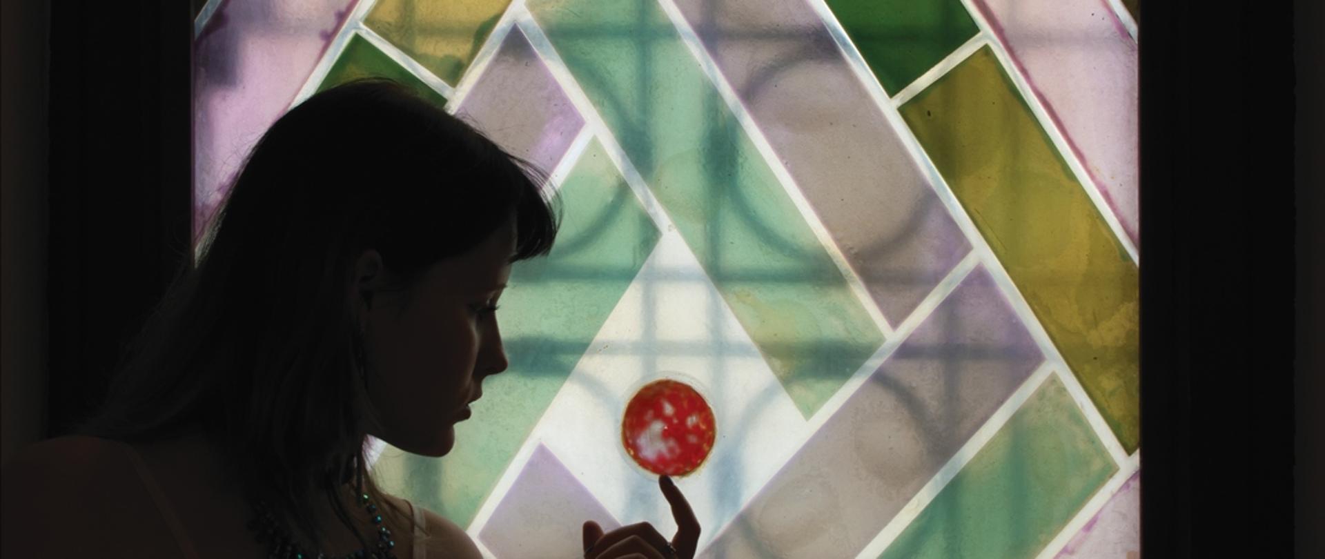 Sylwetka kobiety stojącej przy kolorowym witrażu o geometrycznym wzorze w odcieniach zieleni, różu i żółci; kobieta unosi dłoń i wskazuje palcem mały czerwony, okrągły element na tle jasnego centrum witrażu, całość utrzymana w nastrojowym półmroku.

