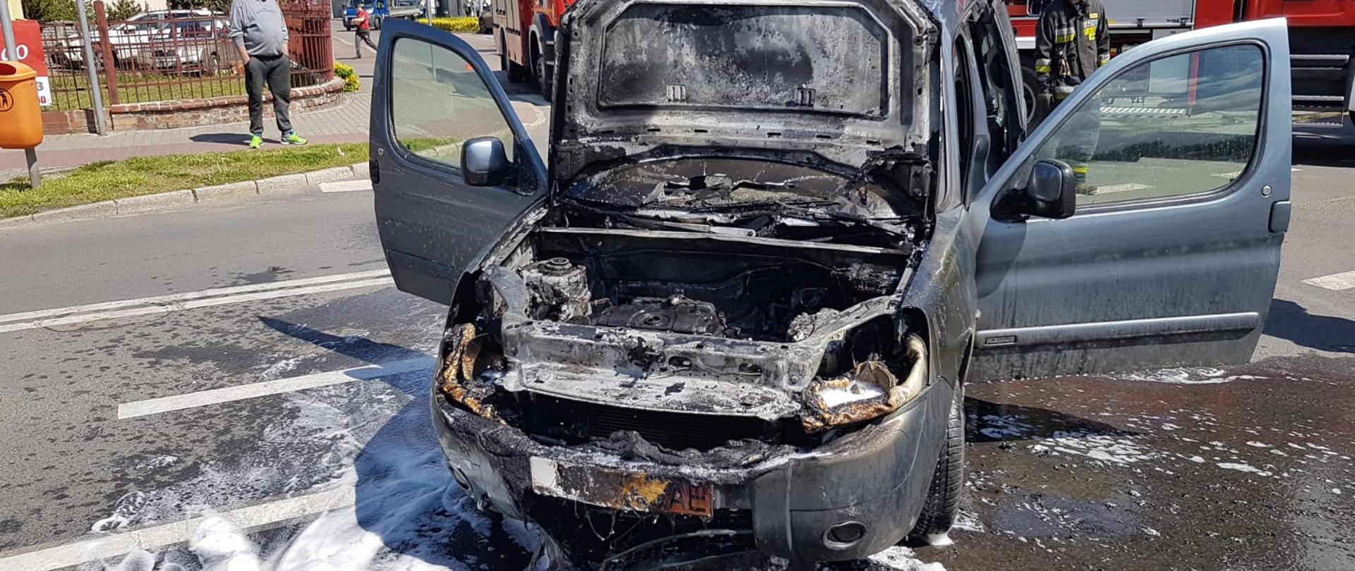 Na zdjęciu spalony samochód stoi na jezdni. Wokół piana gaśnicza. W tle samochód pożarniczy i strażacy.