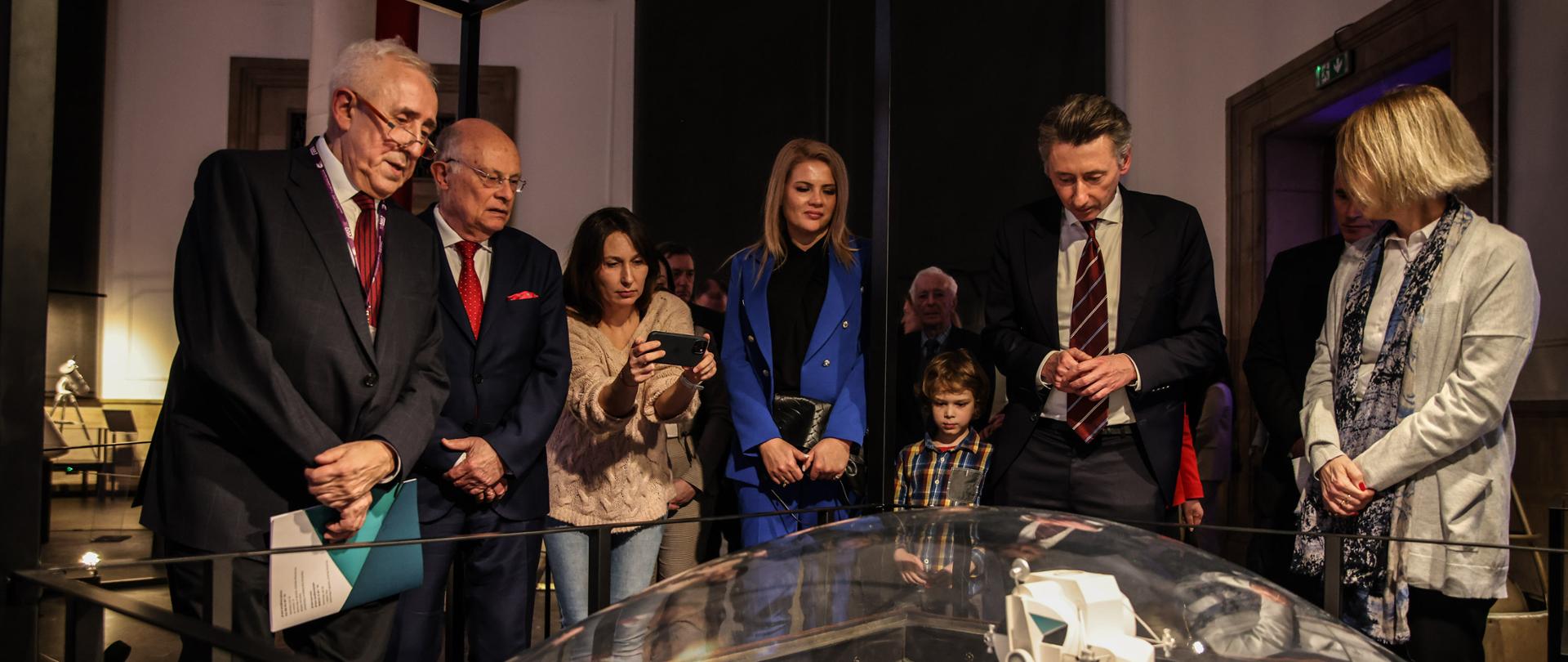 Kilka osób stoi nad zasłoniętą szklaną kopułką makietą przedstawiająca lądownik na powierzchni Księżyca.