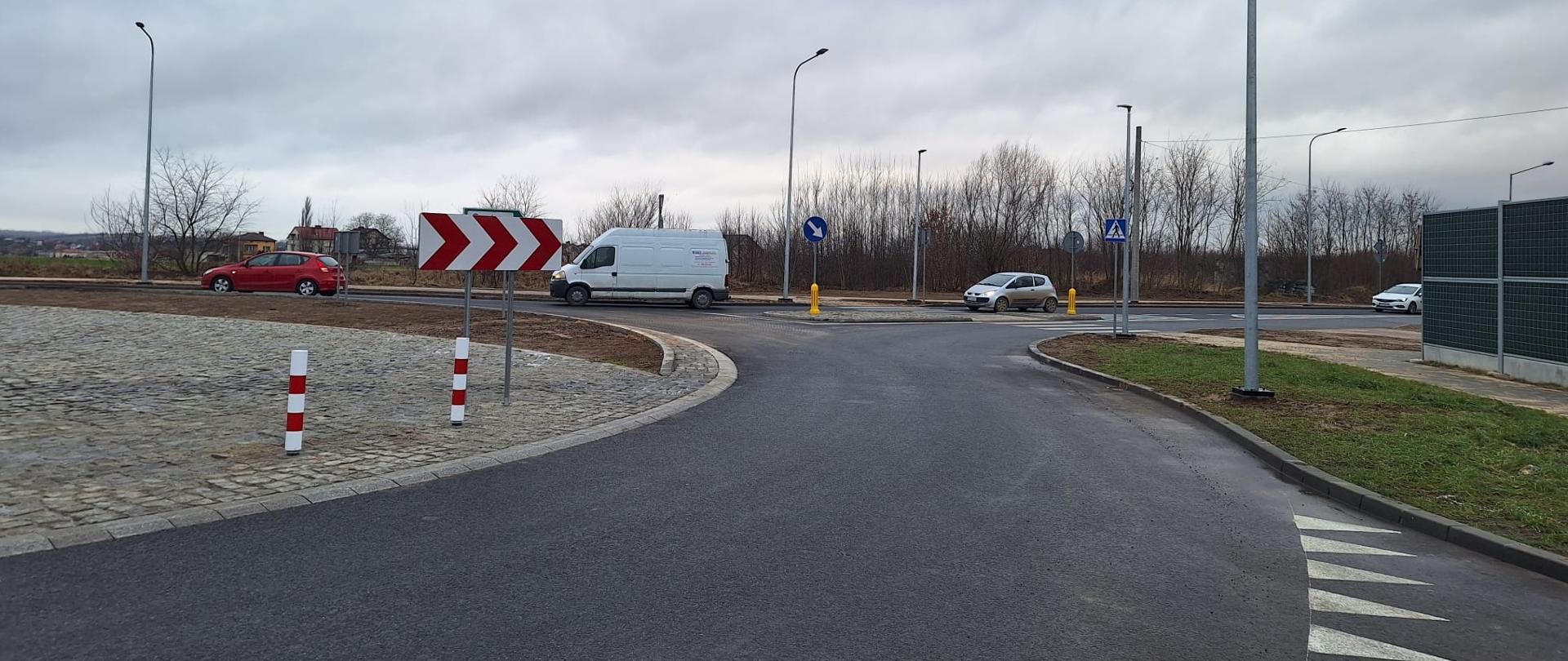 DK9 Ostrowiec Świętokrzyski - rondo na skrzyżowaniu budowanej obwodnicy z istniejącą drogą krajową udostępnione do ruchu. Fragment ronda z nawierzchnią. Na wyspie ronda kostka brukowa, biało-czerwone słupki i oznakowanie. Cztery samochody wjeżdżają na rondo. Po prawej stronie trawnik, chodnik, ekran. 