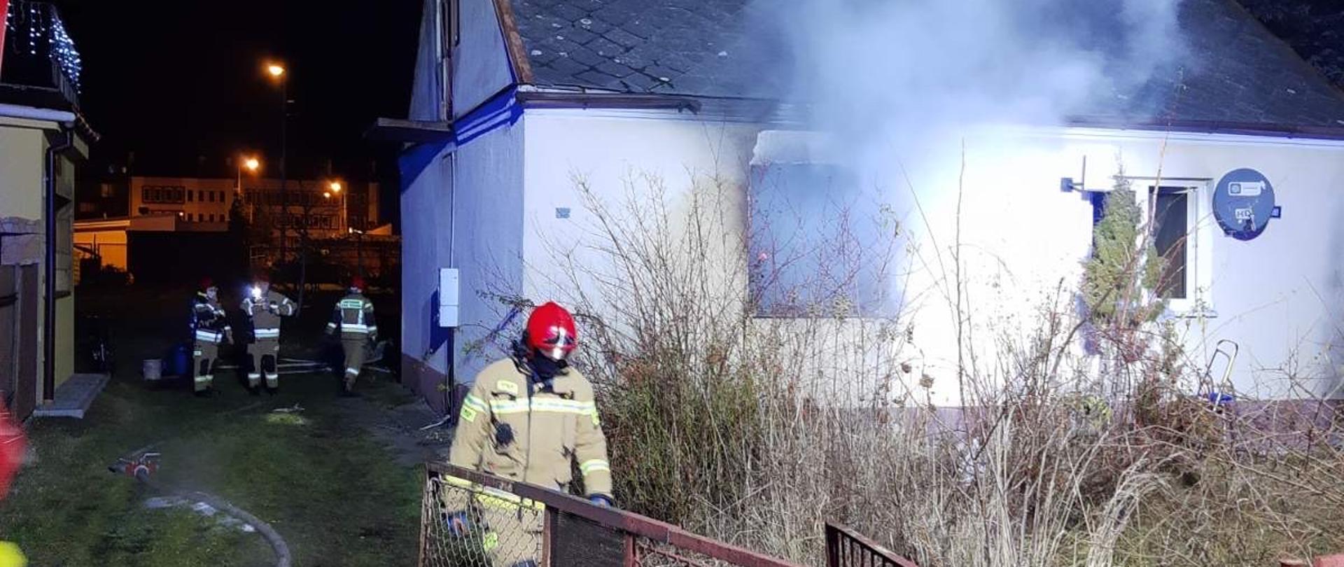 Pora nocna. Budynek mieszkalny, parterowy z dachem dwuspadowym. W ścianie frontowej obiektu dwa oka, z jednego z nich wydobywa się gęsty, biały dym. Przed budynkiem rozłożone na ziemi węże gaśnicze. Na podwórku strażacy. 