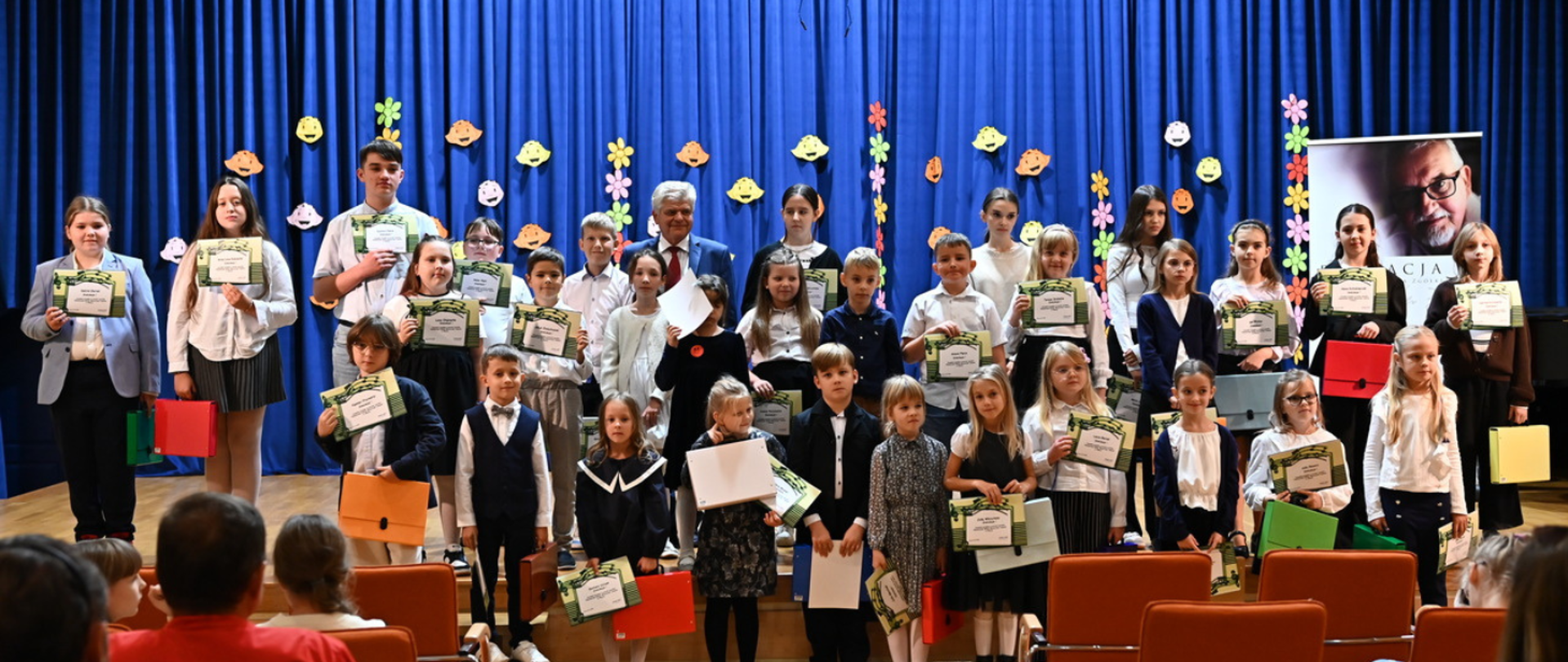 duża grupa uczniów w galowych strojach stojąca na scenie, wszyscy trzymają dyplomy, w tle niebieska kotara