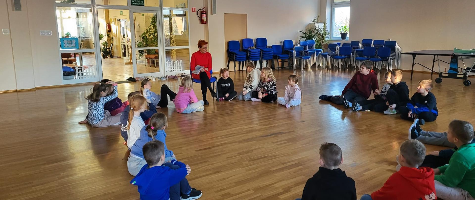 Spotkanie edukacyjne dla dzieci w ramach akcji "Zdrowe ferie". Sala gimnastyczna, dzieci siedzące na podłodze, prelegent siedzi przodem do dzieci.