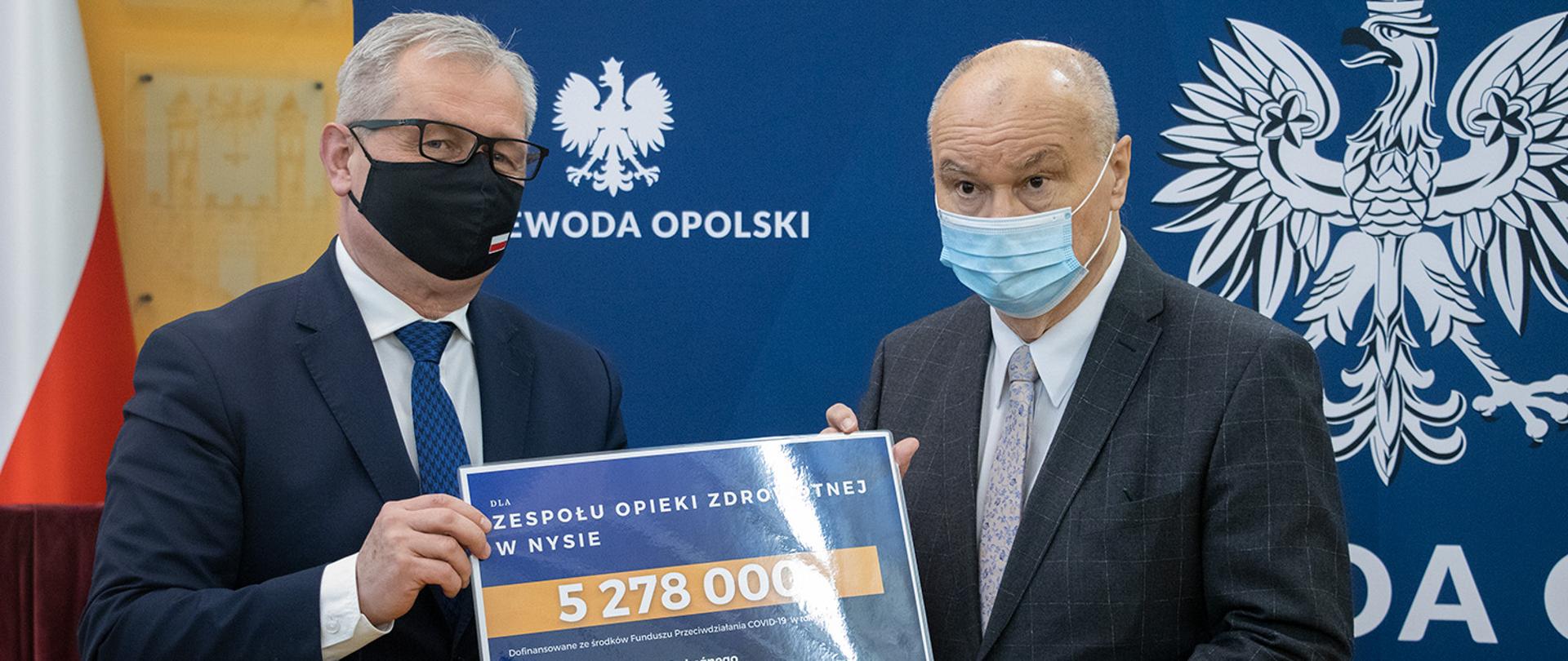 Na zdjęciu wojewoda opolski z dyrektorem nyskiego szpitala, stoją trzymając w dłoniach duzy czek. W tle granatowa ścianka z logo wojewody. 