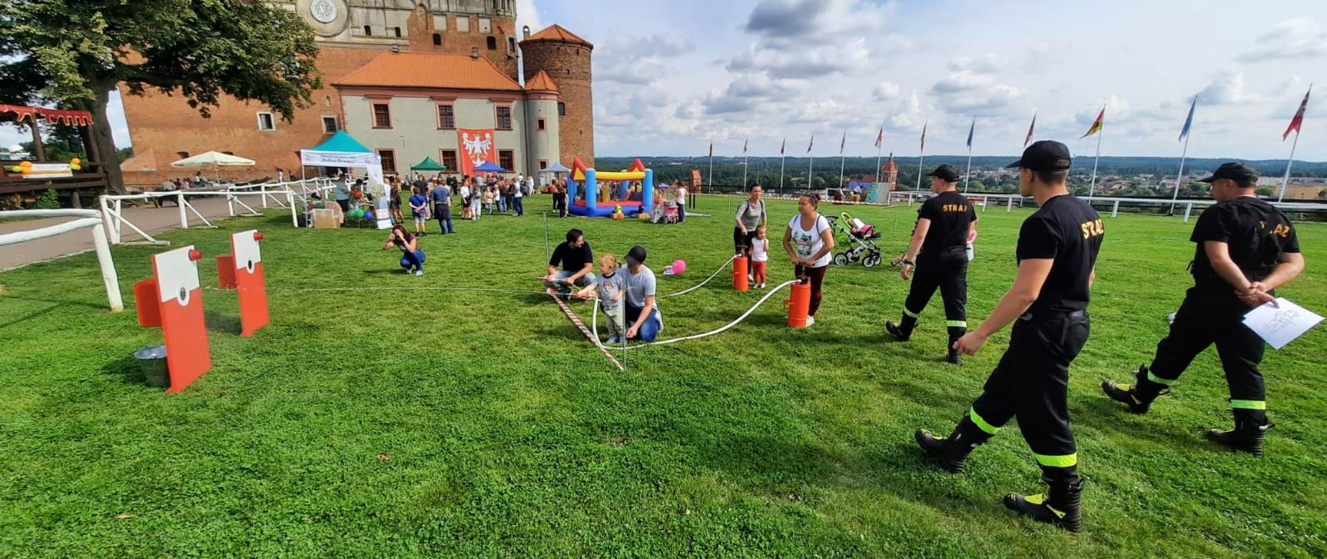 Na zdjęciu widać dzieci oraz osoby dorosłe, które leja wodę do wyznaczonego celu. po prawej stronie widać idących strażaków w ubraniach koszarowych. W tle widać zamek z cegieł.