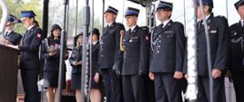 grupa kilkunastu strażaków osp oraz komendant powiatowy psp w Łowiczu ubrani w umundurowanie wyjściowe z czapka rogatywka na głowie oczekują na odznaczenie pamiątkowym medalem, na pierwszym planie przy mównicy kobieta i mężczyzna ubrani w mundury wyjściowe psp, kobieta mówi do mikrofonu