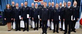 Zdjęcie grupowe - na pierwszym planie mazowiecki komendant wojewódzki PSP, za nim grupa oficerów prasowych. Wszyscy występują w mundurach wyjściowych. Nagrodzeni trzymają w rękach białe torby i trzymają białe teczki. Za nimi niebieskie tło oraz wiszące godło państwowe.