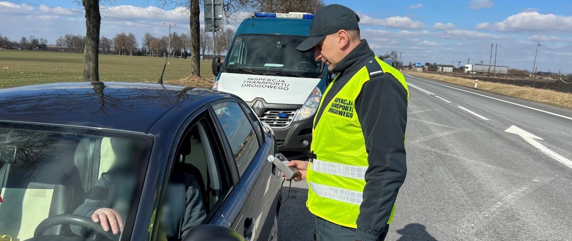 Na pierwszym planie stoi inspektor świętokrzyskiej ITD obok zatrzymanego samochodu osobowego i trzyma w ręku alkomat. W tle oznakowany radiowóz ITD typu furgon.