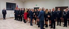 W pomieszczeniu o jasnych ścianach i podłodze strażacy stoją ustawieni jeden obok drugiego. Kobieta i dwóch mężczyzn w garniturach składają im gratulacje. Za strażakami widać sztandar. Z lewej strony na tel jasnej ściany z oknami stoi strażak w postawie na baczność.
