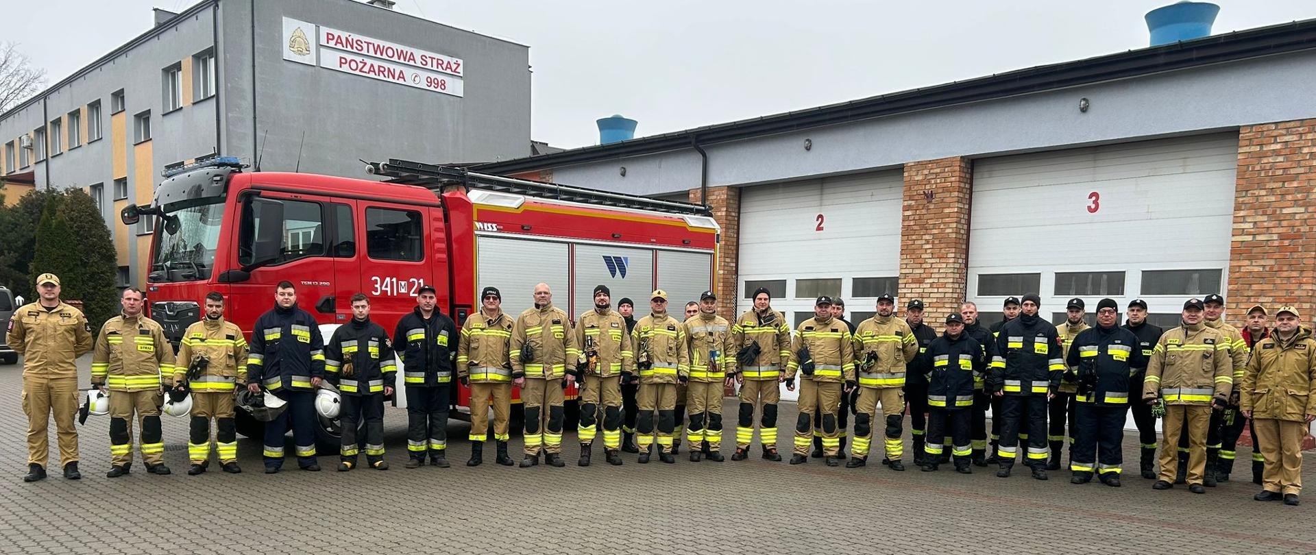 Na zdjęciu widać dużą grupę strażaków stojących w szeregu przed budynkiem Komendy Miejskiej PSP w Ostrołęce. Za nimi znajdują się zamknięte bramy garażowe oznaczone numerami „2” i „3”, a z lewej strony widoczny jest czerwony samochód ratowniczo-gaśniczy z oznaczeniem „341[M]21”. Strażacy są ubrani w różne zestawy umundurowania bojowego: część w piaskowe ubrania specjalne, część w granatowe oraz spodnie z odblaskami. Wszyscy stoją równo, pozując do zdjęcia grupowego przed budynkiem strażnicy po egzaminie szkolenia kierowcy-konserwatora sprzętu ratowniczego OSP.