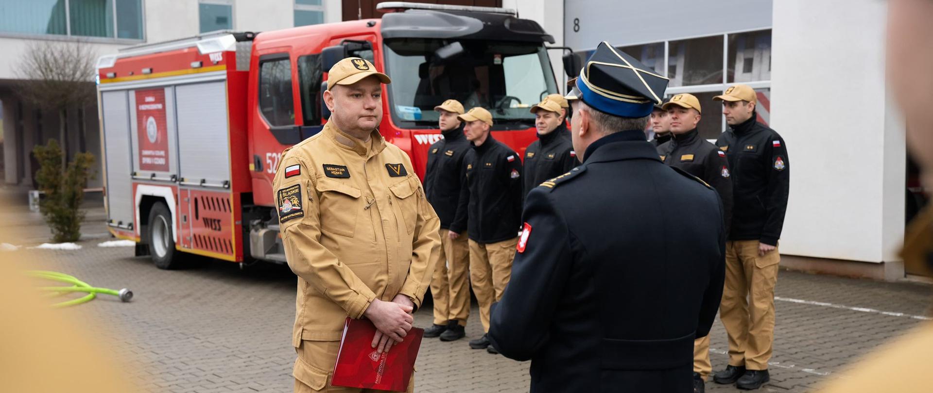 Na zdjęciu widać uroczystą zdanie służby przed budynkiem straży pożarnej. Na pierwszym planie stoi strażak przechodzący na emeryturę. Naprzeciwko niego stoi komendant, galowym płaszczu Jest odwrócony tyłem do obiektywu i wręcza życzenia. W tle ustawionych jest kilku innych strażaków w jednakowych mundurach. Za nimi zaparkowany jest czerwony wóz strażacki 
