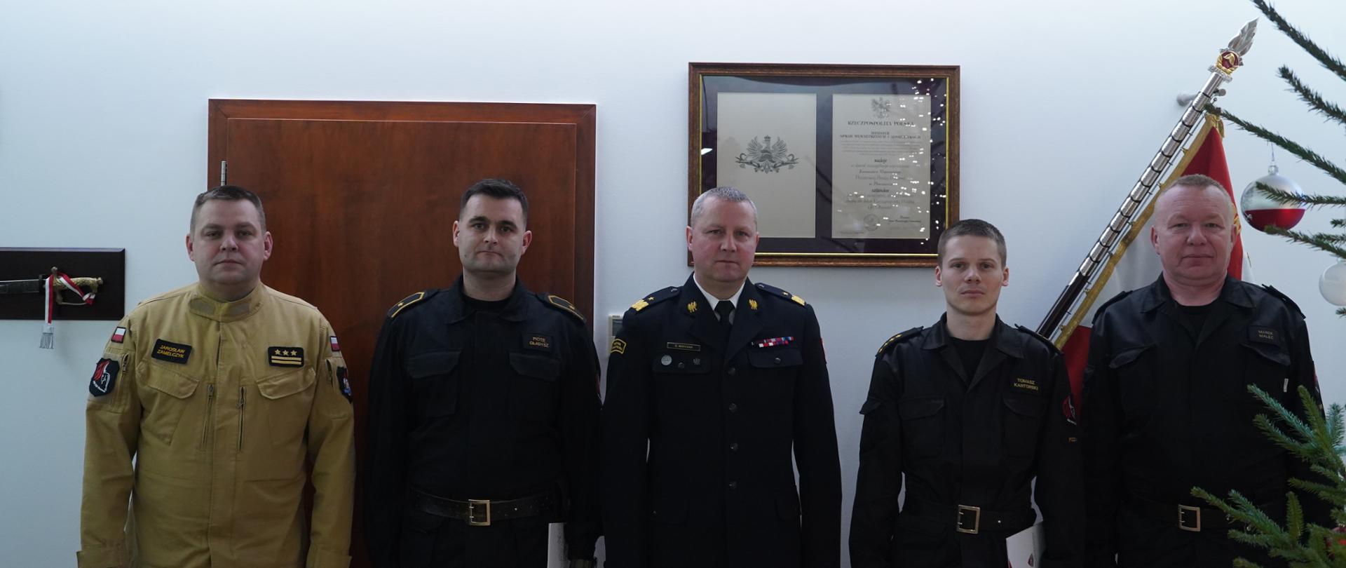 pięciu strażaków stoi obok siebie, w środku strażak w mudurze wyjściowym - marynarka i spodnie, z prawej strony dwóch strażaków w czarnym mundurze dowódczo-sztabowym, z lewej jeden strażak w czarnym mundurze i obok jeden w jasnym miodowym mundurze, w tle sztandar 