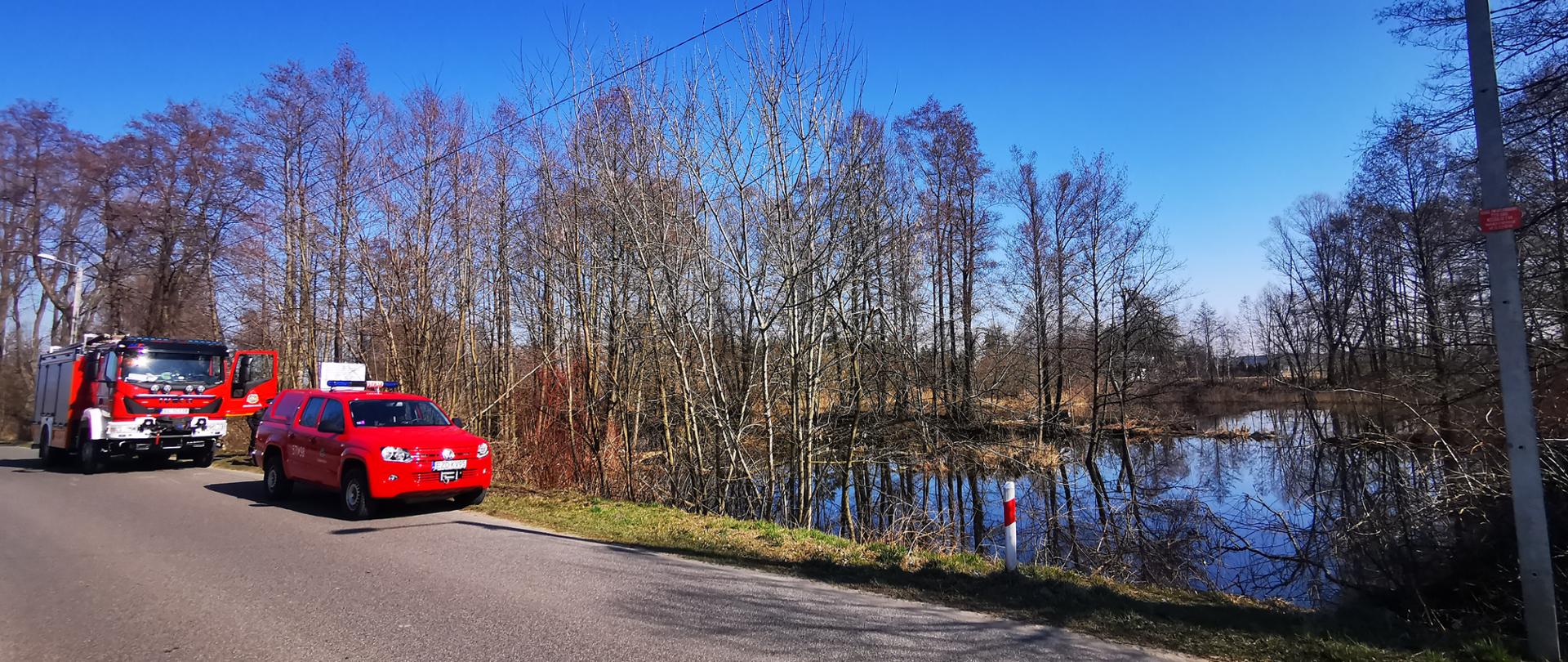 Na zdjęciu widoczny jest staw przy utwardzonej drodze gminnej, na poboczu zaparkowane są czerwone samochody pożarnicze. 