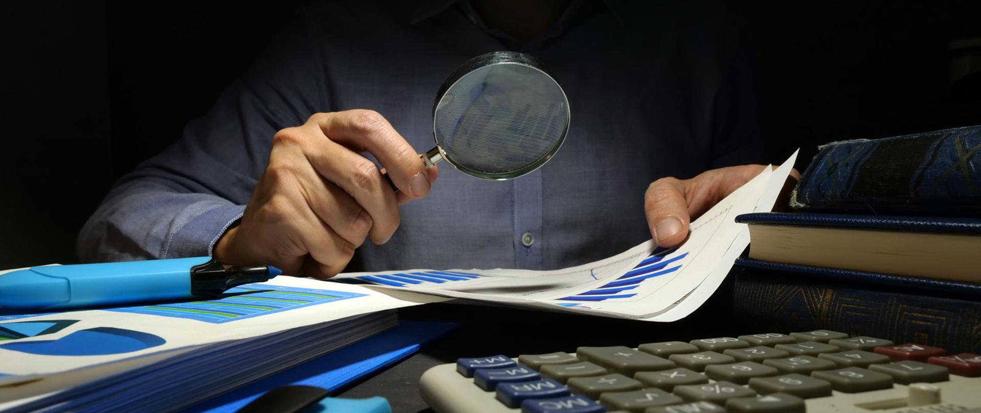 Dłoń trzymająca lupę nad dokumentami, kalkulator.